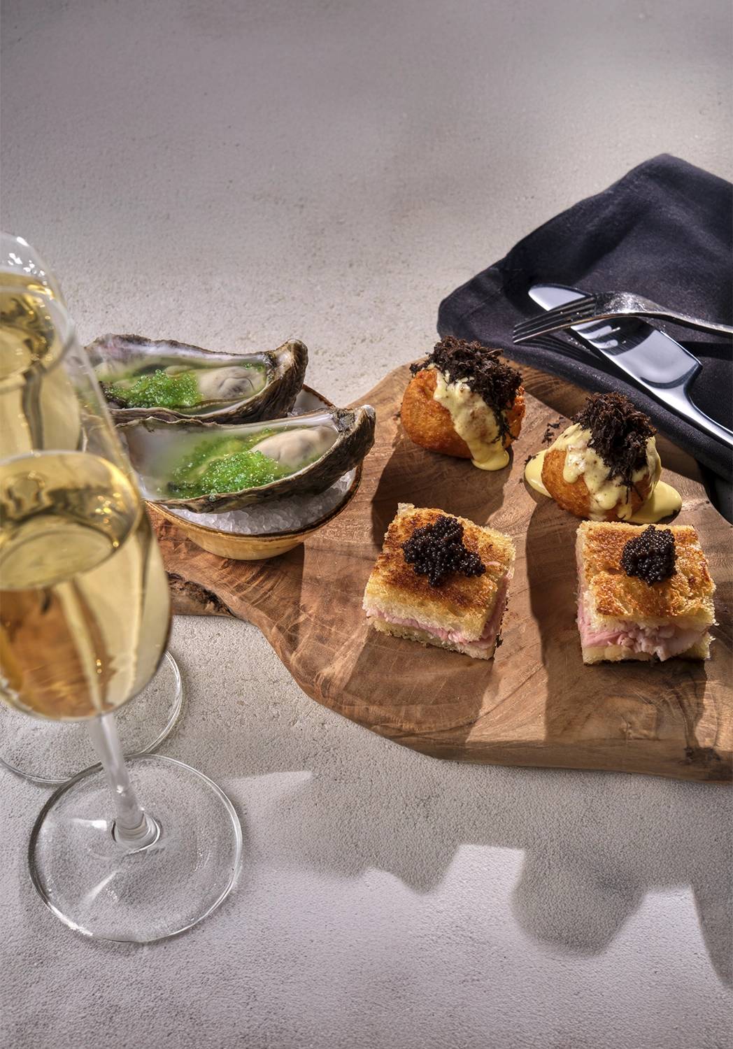 Wooden board with gourmet canapés including oysters with green garnish, fried bites with creamy sauce and black topping, and small sandwiches topped with black caviar, accompanied by two glasses of champagne and cutlery on a black napkin.