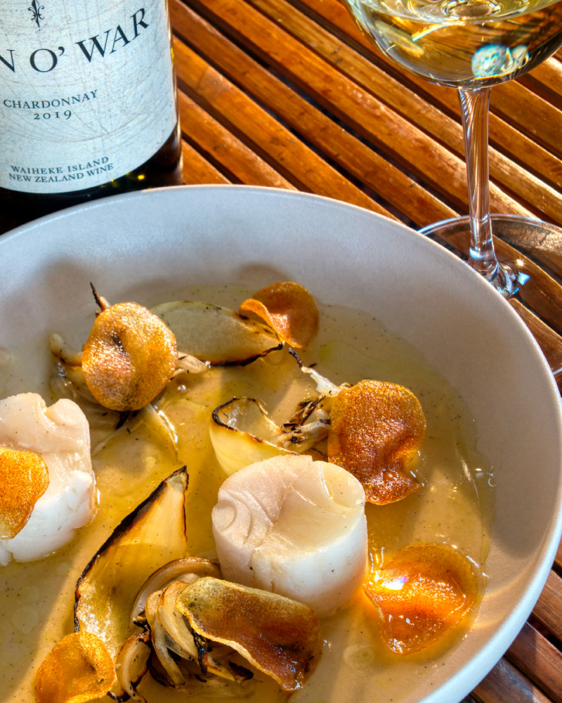 Bowl of scallops with grilled onions and crispy chips, served with a glass of white wine and a bottle of Chardonnay.
