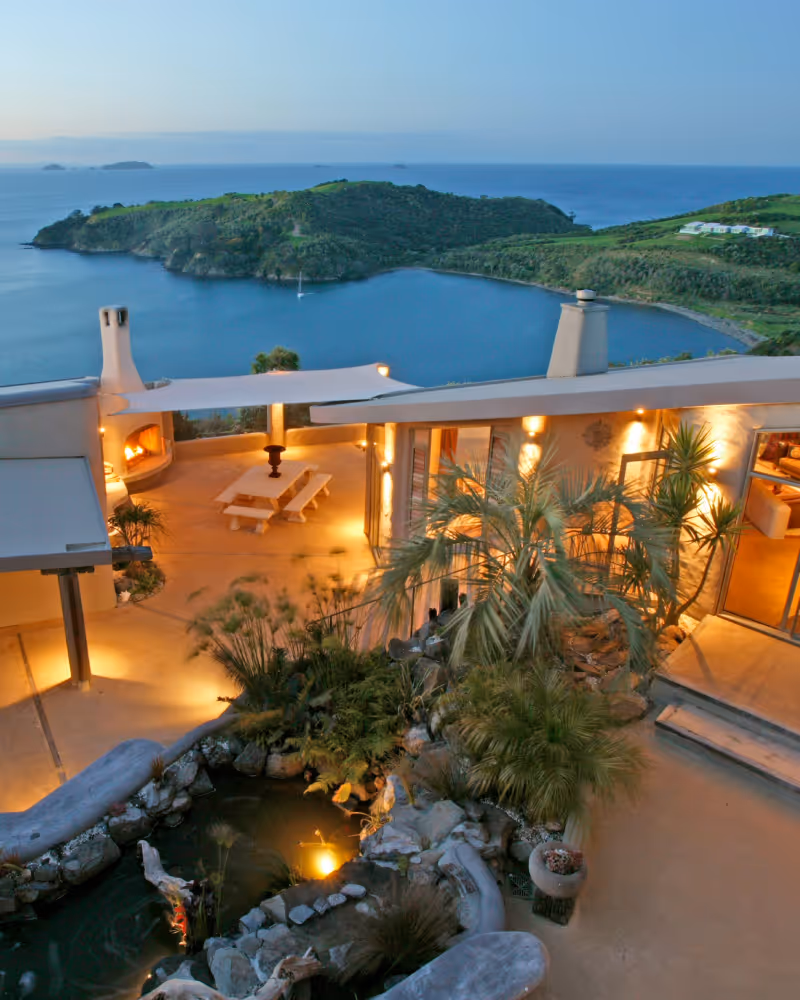 Evening view of a lit outdoor patio with fireplace, picnic table, lush plants, and a bay with green hills in the background.