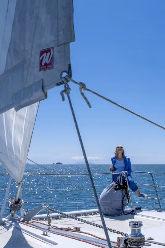 Woman in blue outfit sitting on the deck of a sailboat under clear blue sky and calm sea.
