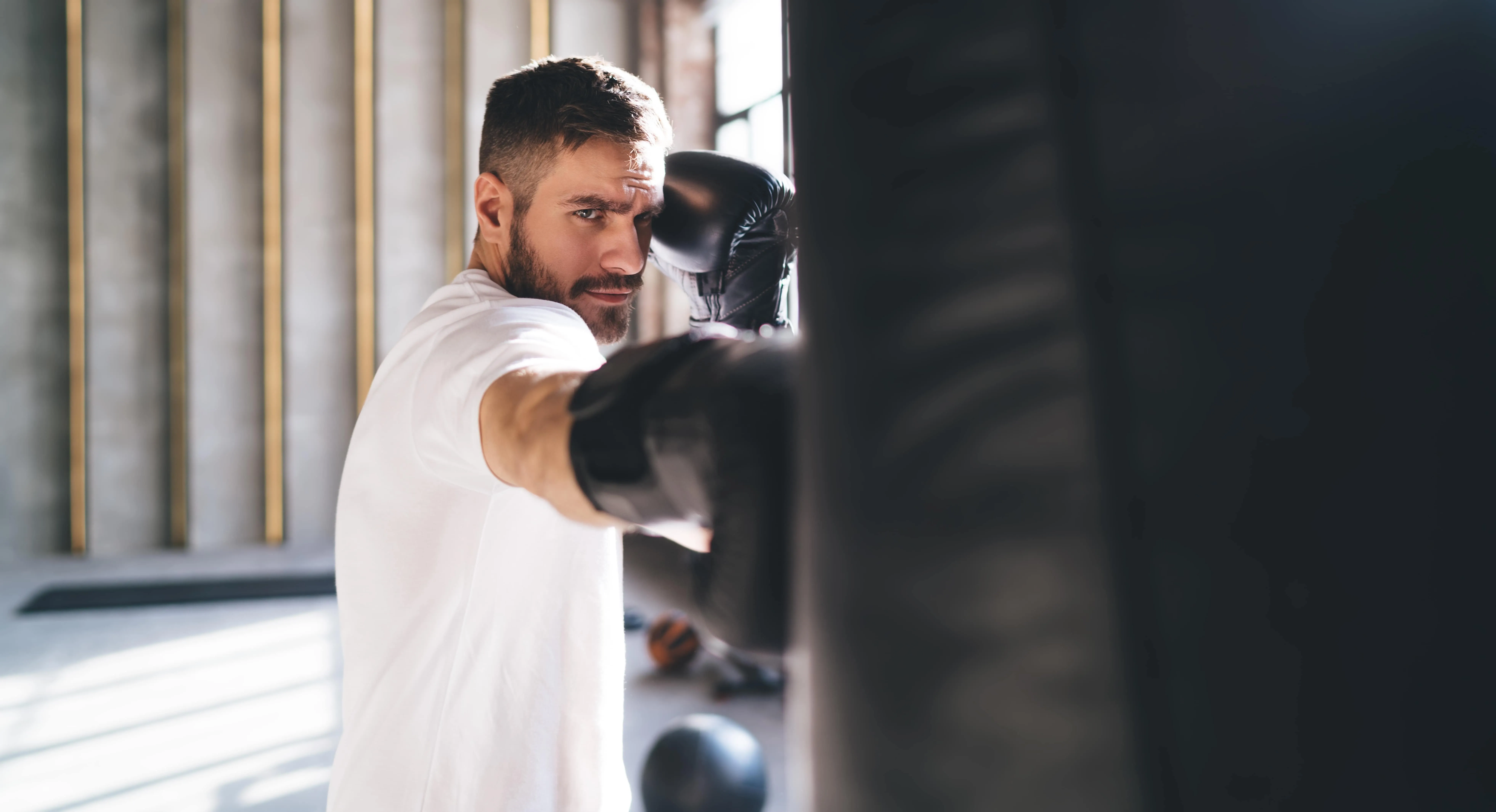 Man in white shirt wearing black boxing gloves punching a heavy bag in a sunlit gym.