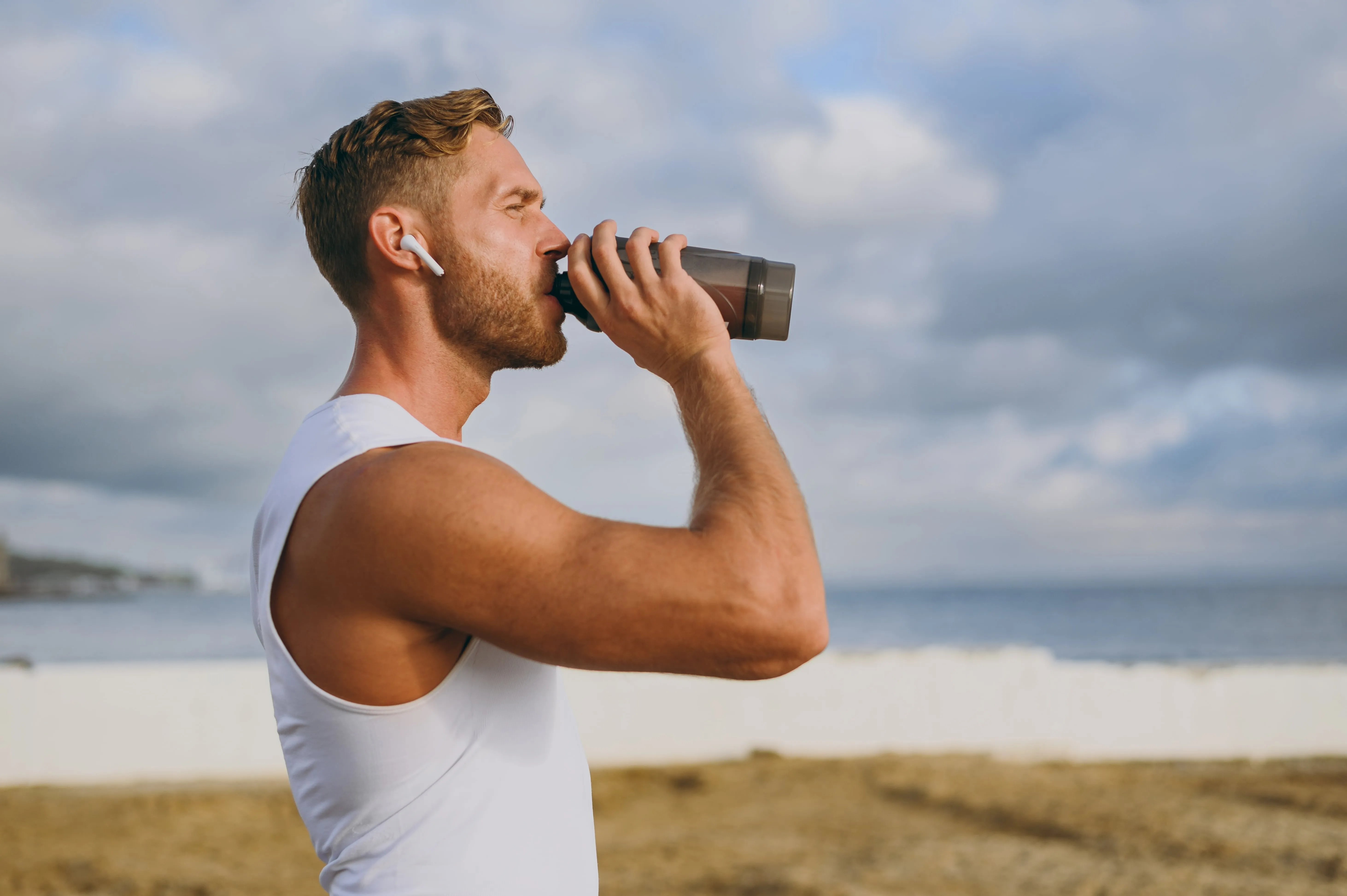 Man wearing white tank top and wireless earbuds drinking from a black shaker bottle outdoors near a body of water.