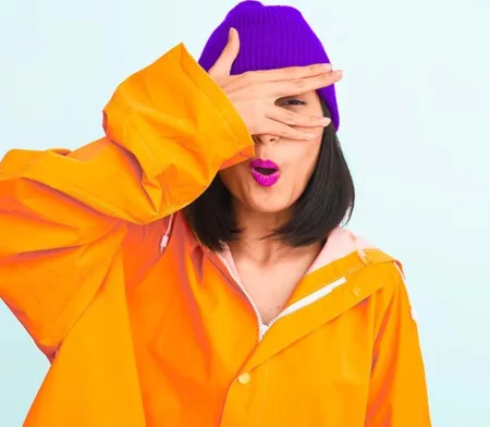 mujer con chaqueta naranja y gorro morado mirando entre los dedos