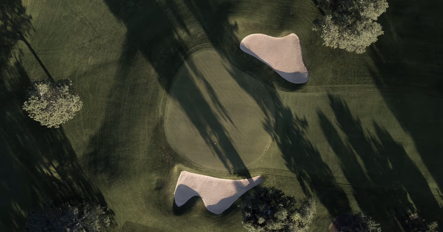 Aerial view of a golf green with two sand bunkers and surrounding trees casting long shadows.