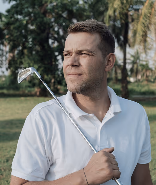Man in white polo shirt holding a golf club over his shoulder and looking into the distance in a green outdoor setting.