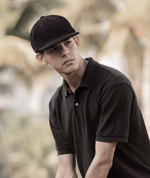 Young man wearing a black cap and black polo shirt looking off to the side against a blurred outdoor background.