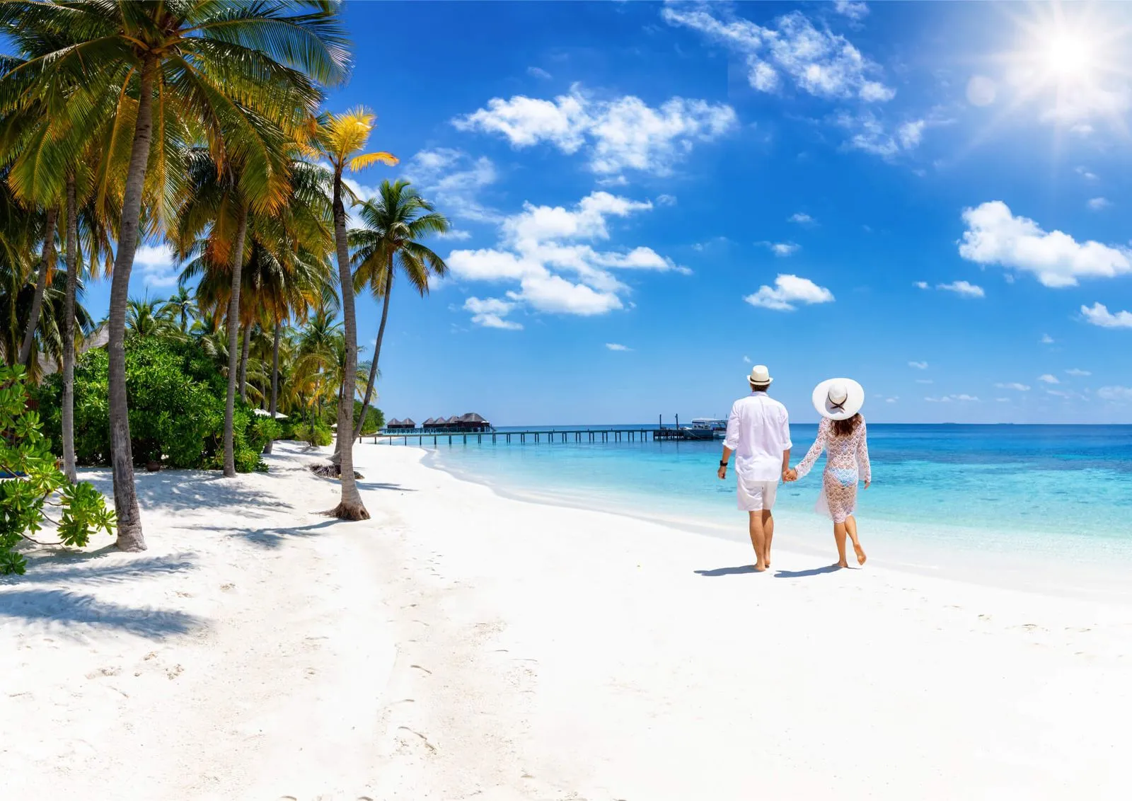 Couple holding hands walking barefoot on a sunny white sand beach with clear blue ocean.