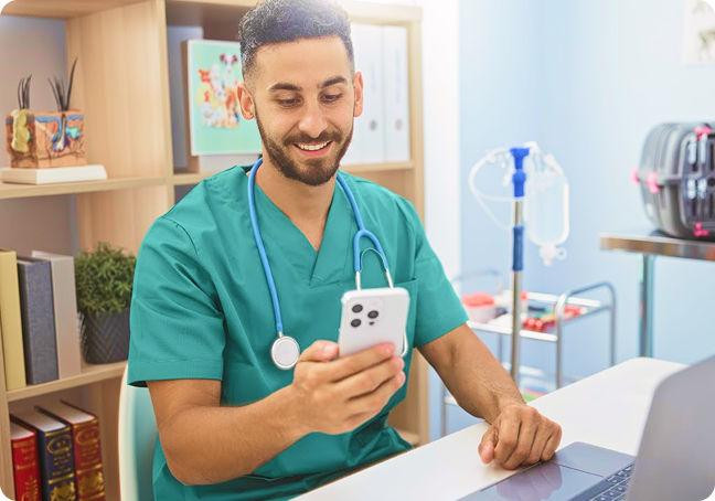 Smiling male healthcare worker in scrubs looks at smartphone while sitting at a desk with a laptop.