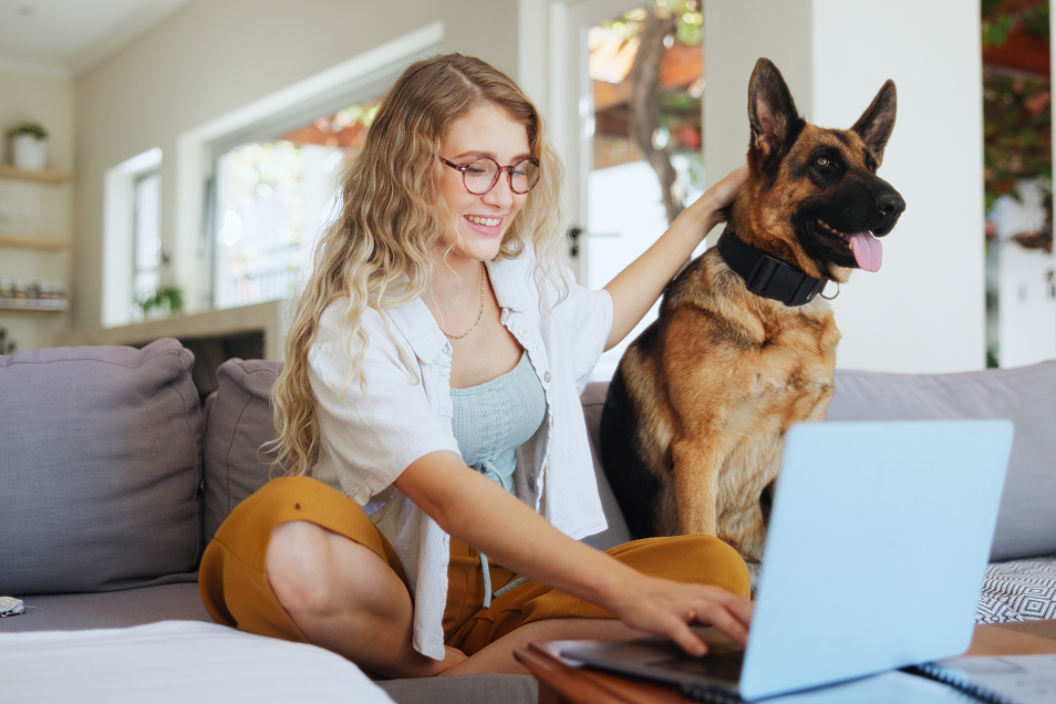 Smiling woman with glasses using laptop and petting German shepherd sitting beside her on couch.