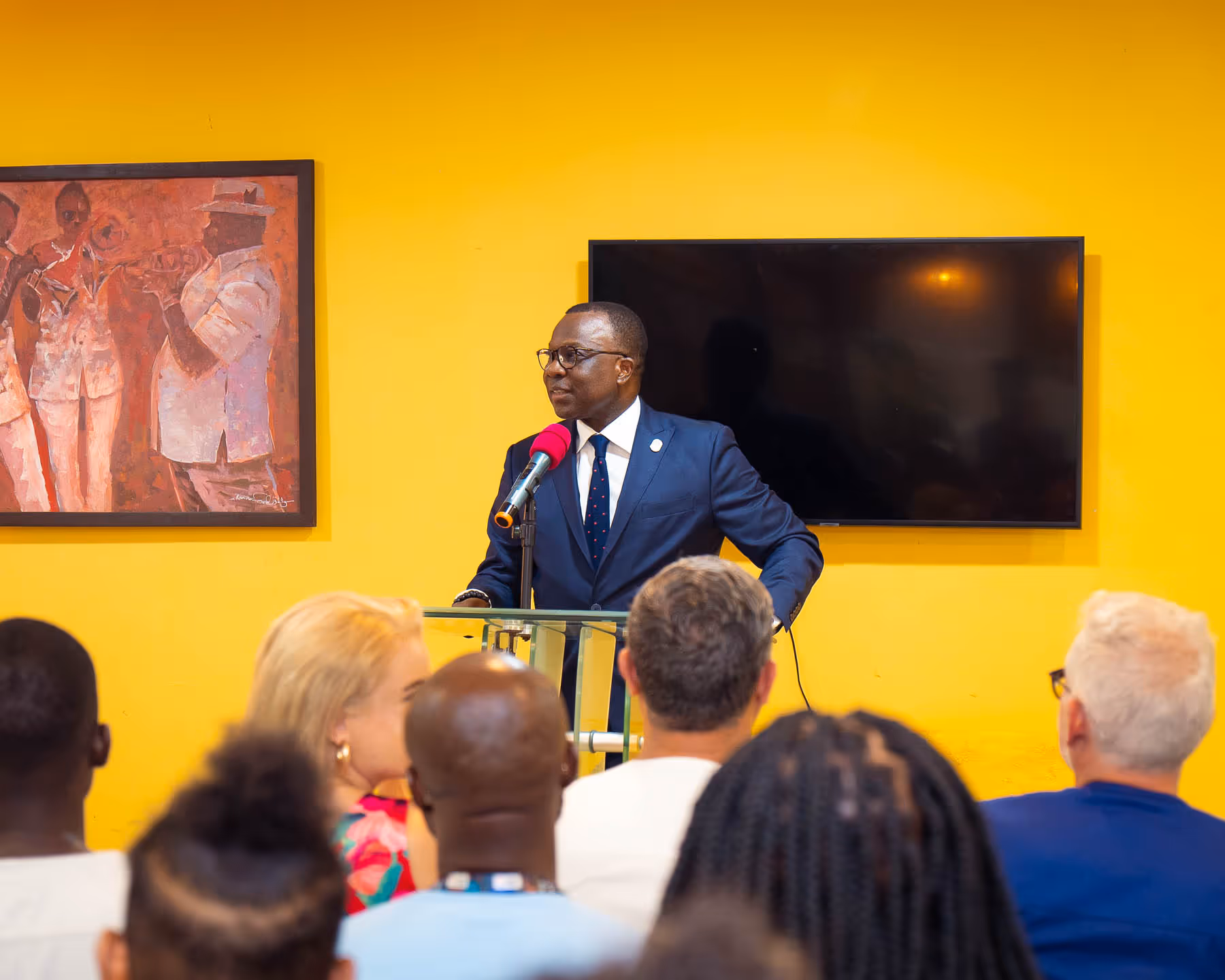 Man in a navy suit speaking at a podium with a microphone in front of a yellow wall with a TV screen and a framed painting.