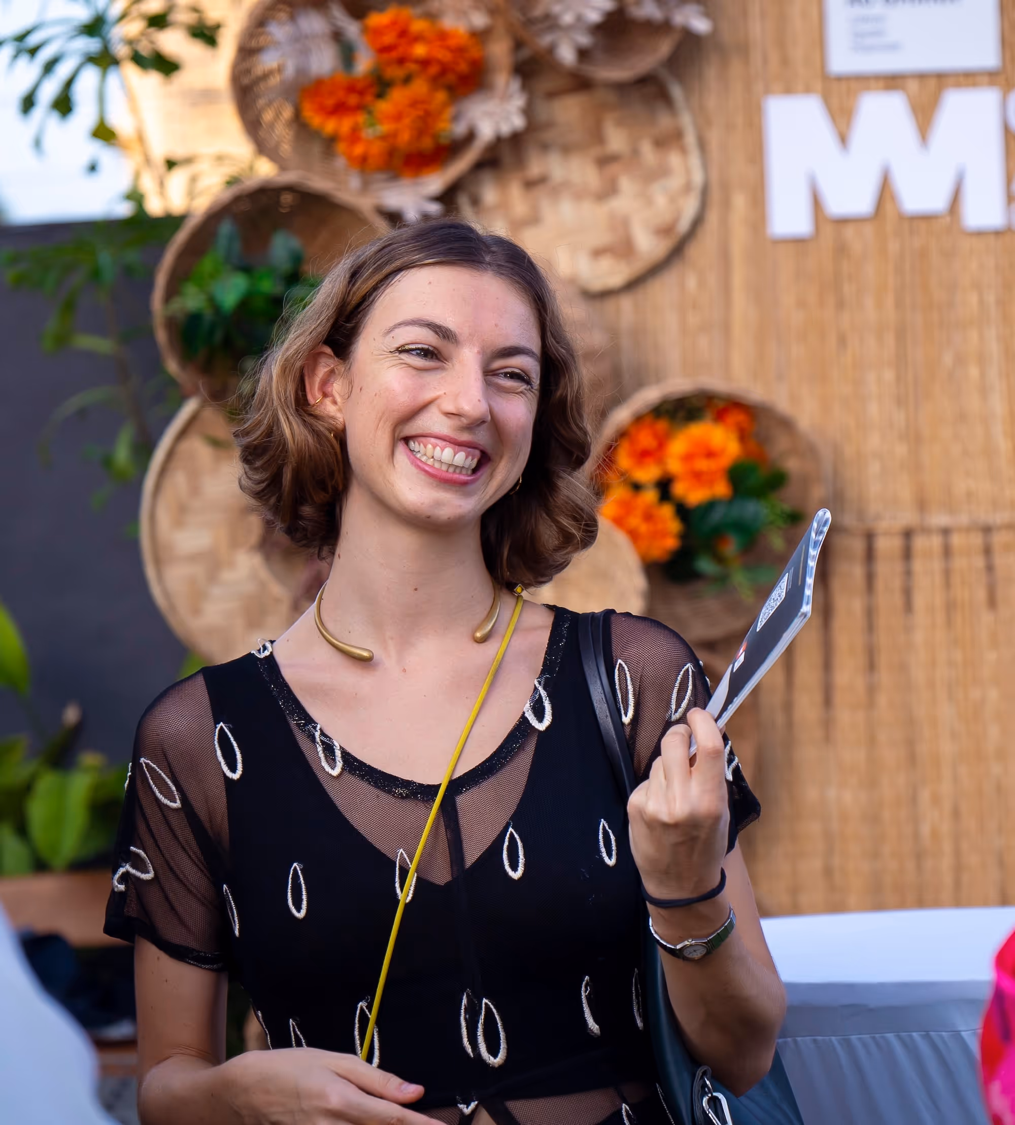 Smiling woman with short brown hair wearing a black patterned sheer top, holding a card in one hand.
