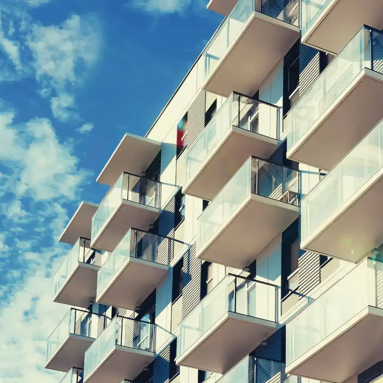 Façade d'un immeuble moderne avec plusieurs balcons en verre sous un ciel bleu partiellement nuageux.