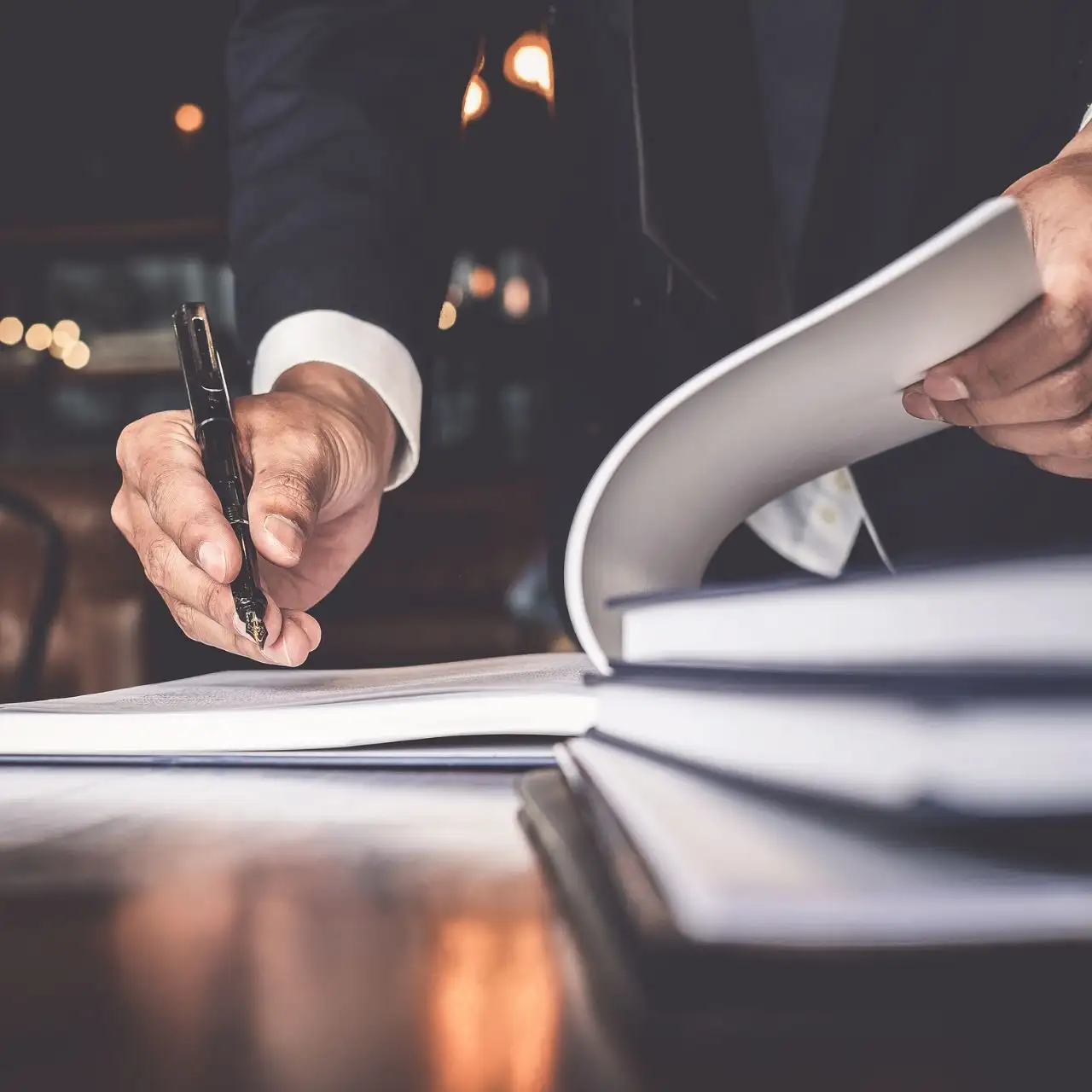 Main d'une personne en costume tenant un stylo et feuilletant des pages dans un grand livre ouvert sur une table en bois.