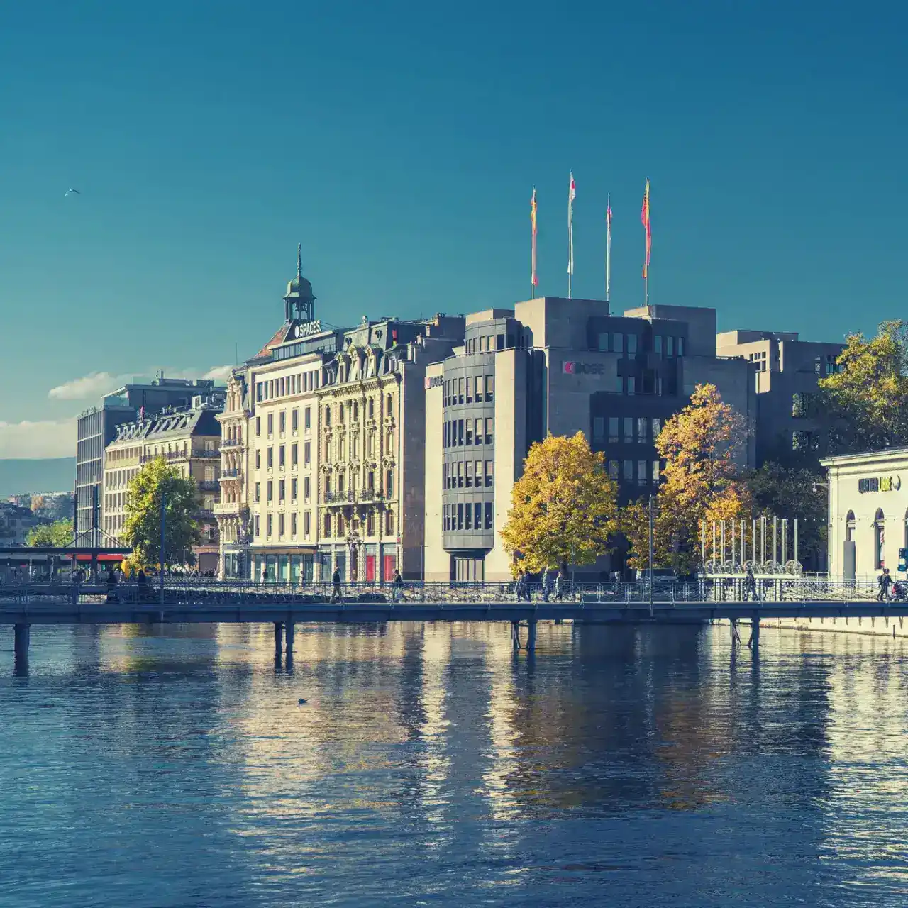 Bâtiments historiques et modernes au bord d’une rivière avec un pont piétonnier et des arbres aux couleurs automnales sous un ciel bleu.