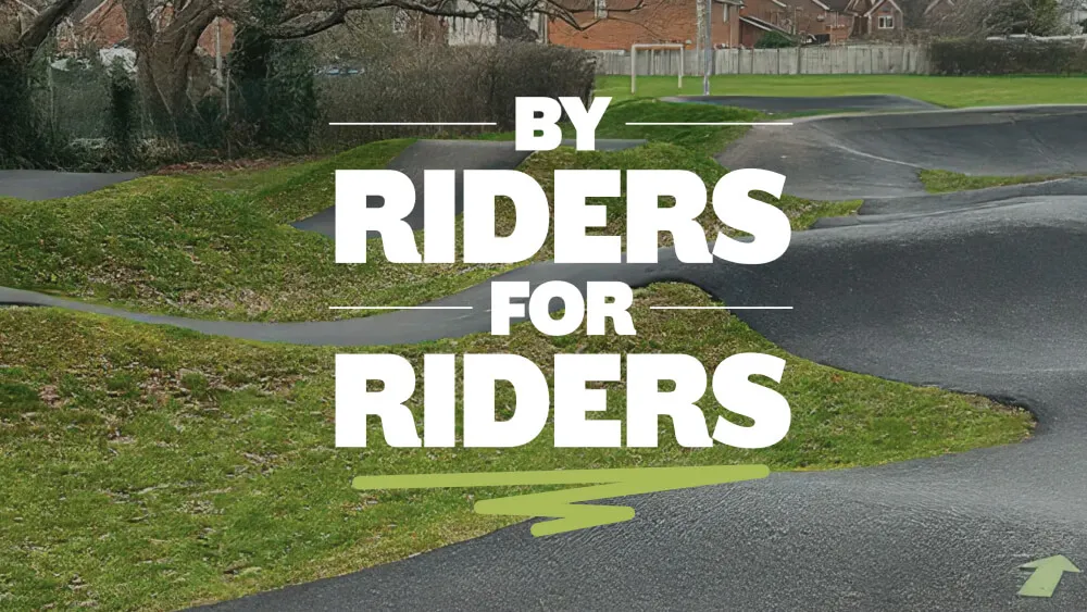 Outdoor pump track with paved winding paths and grassy mounds featuring the text 'BY RIDERS FOR RIDERS' in bold white letters.