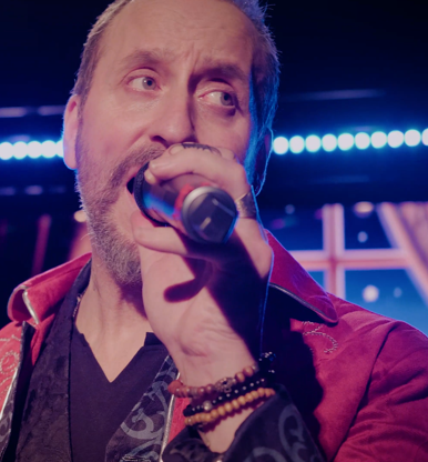 Man with a beard singing into a microphone on stage with blue lights in the background.