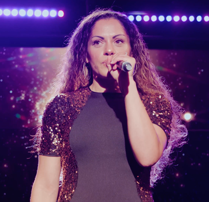 Woman with long curly hair singing into a microphone on stage with bright purple lights in the background.