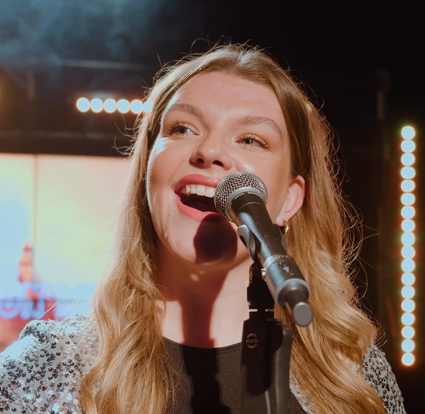 Young woman with long blonde hair smiling and singing into a microphone on stage with bright lights in the background.