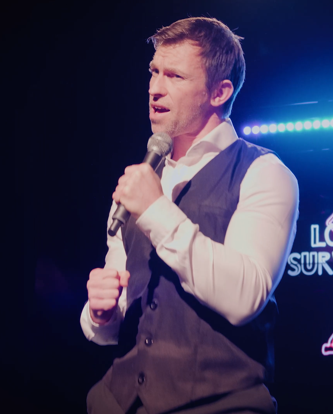 Man holding a microphone, speaking on stage with blue lighting in the background.