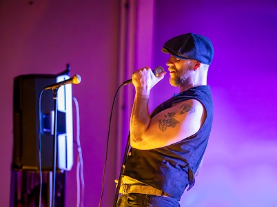 Man with a flat cap and sleeveless vest singing into a microphone on stage with purple lighting.