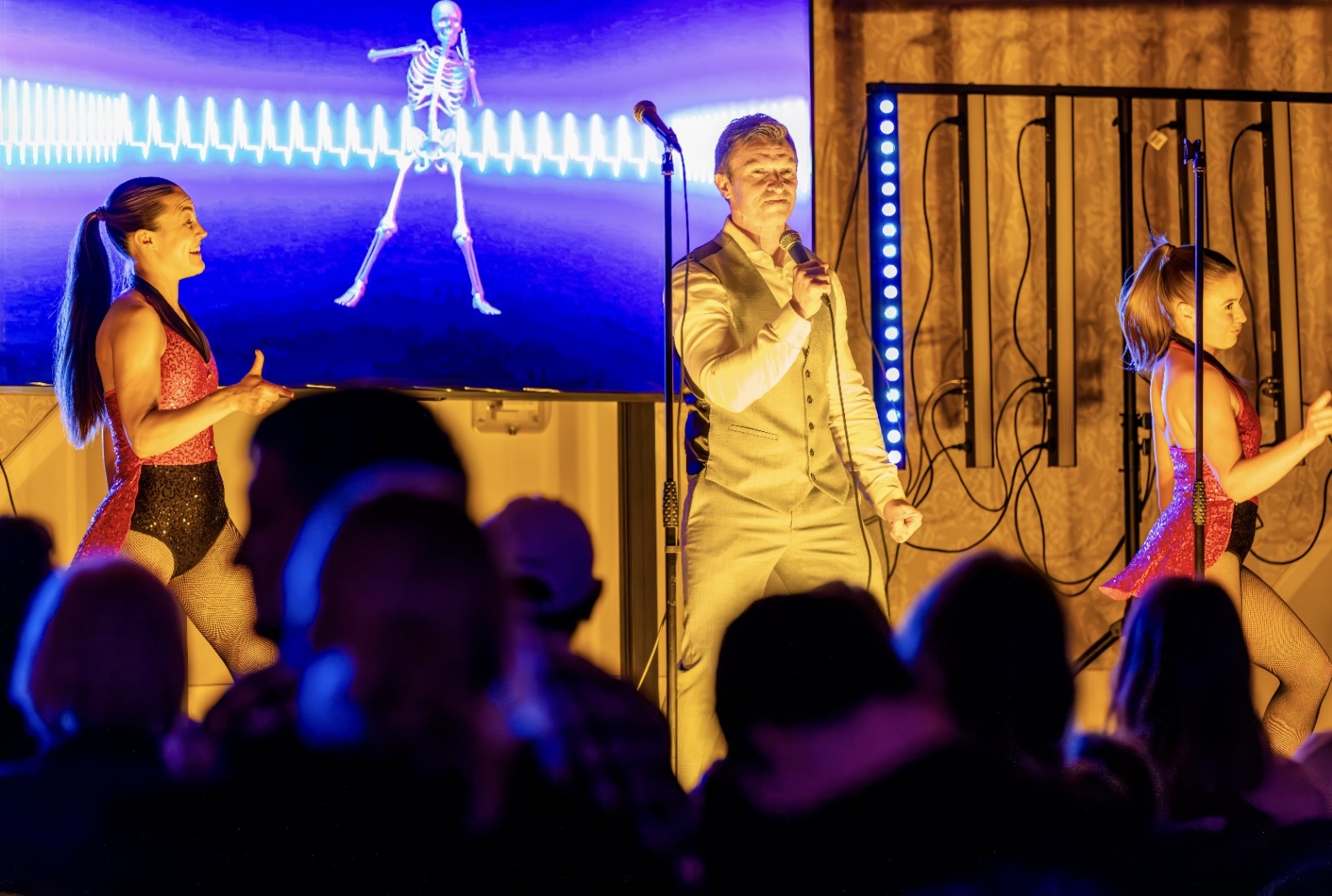 A man in a light suit sings into a microphone on stage with two women dancers in red outfits and a screen behind showing a dancing skeleton graphic.