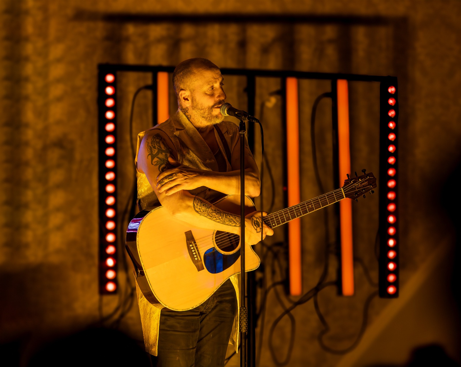 Tattooed male musician singing into a microphone while holding an acoustic guitar on stage with orange lights in the background.