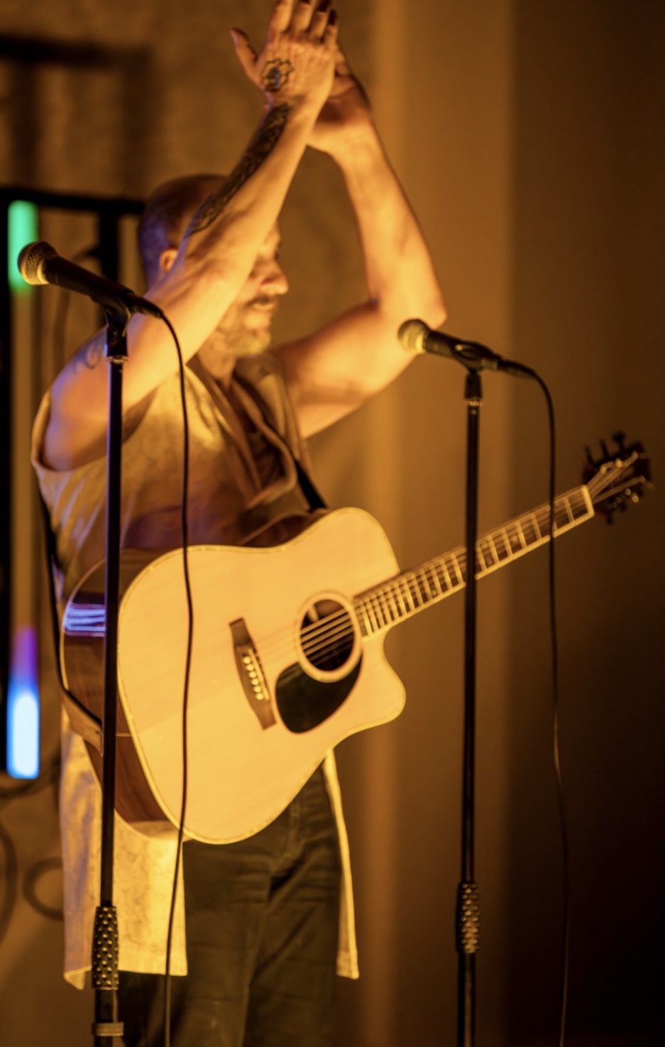 Musician with tattoos clapping hands above his head while holding an acoustic guitar on stage with two microphones.
