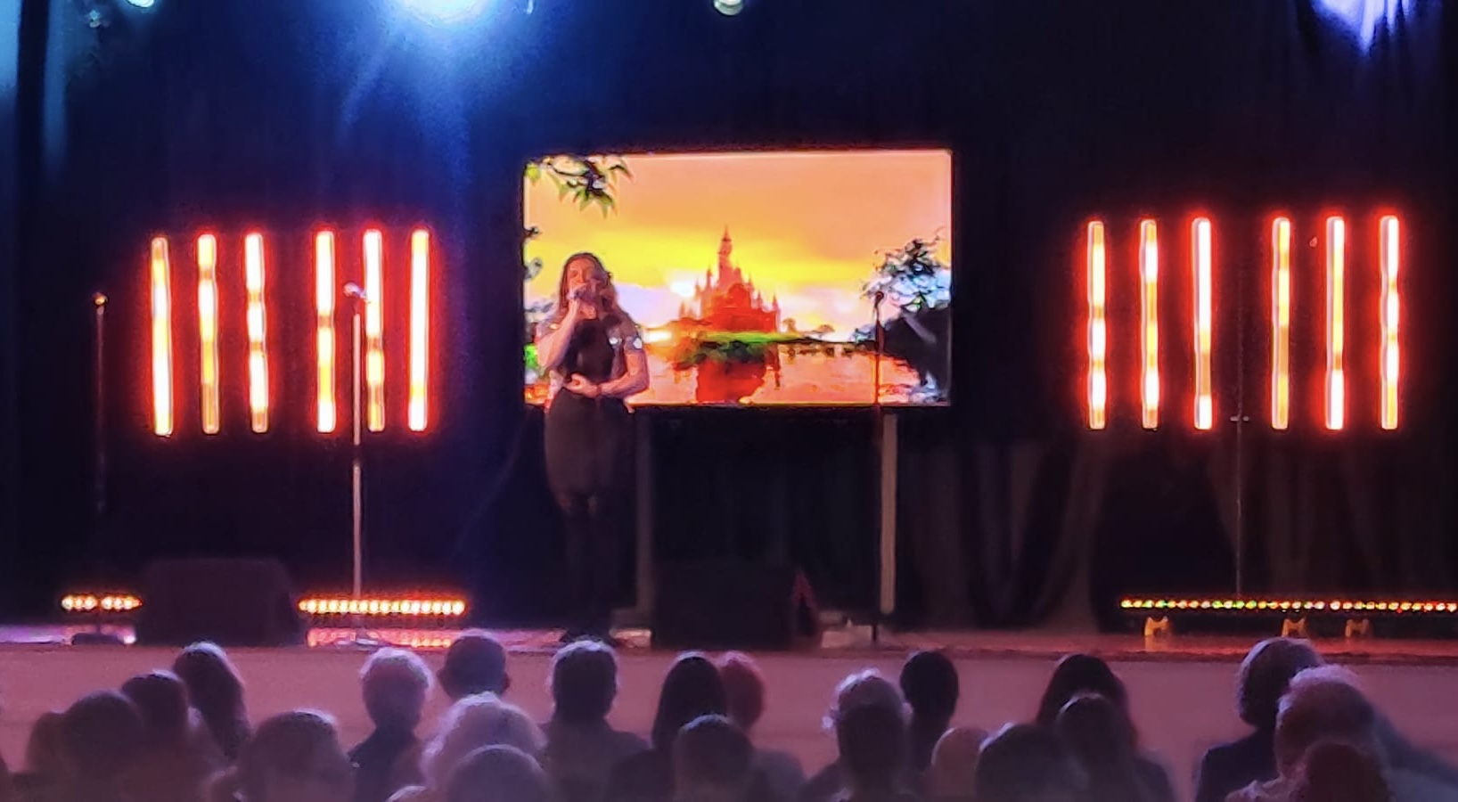 A man in a light suit sings into a microphone on stage with two women dancers in red outfits and a screen behind showing a dancing skeleton graphic.