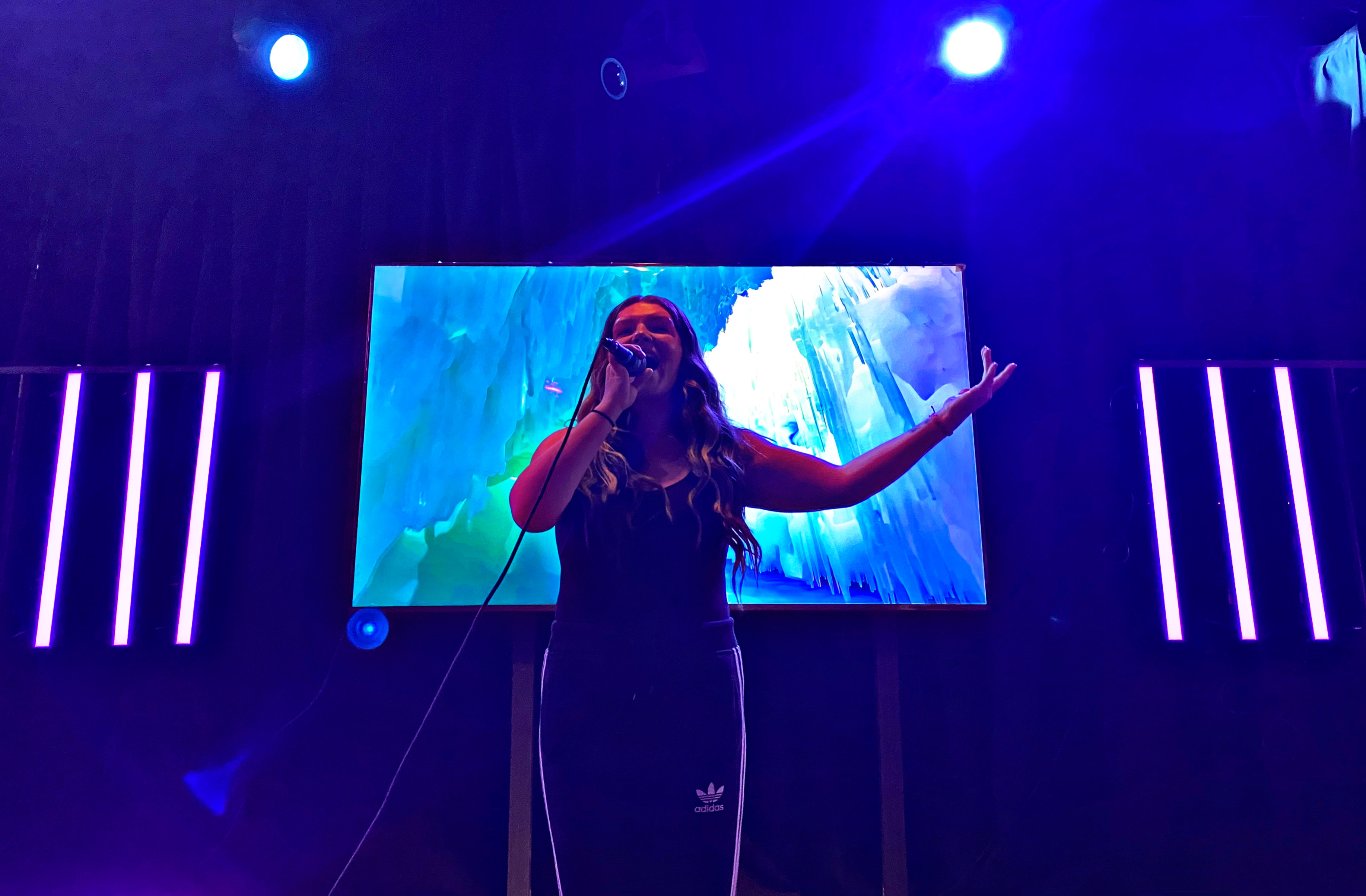 A man in a light suit sings into a microphone on stage with two women dancers in red outfits and a screen behind showing a dancing skeleton graphic.