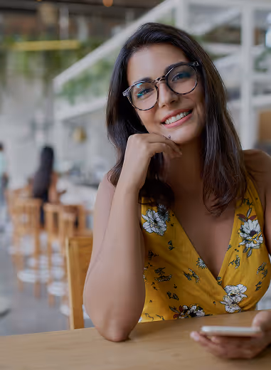 Smiling woman with glasses in a yellow floral dress sitting at a wooden table holding a phone in a bright cafe.