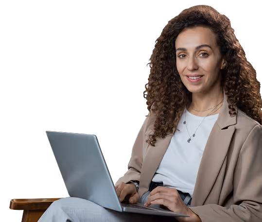 Young woman with curly hair sitting on a wooden chair using a laptop and smiling.