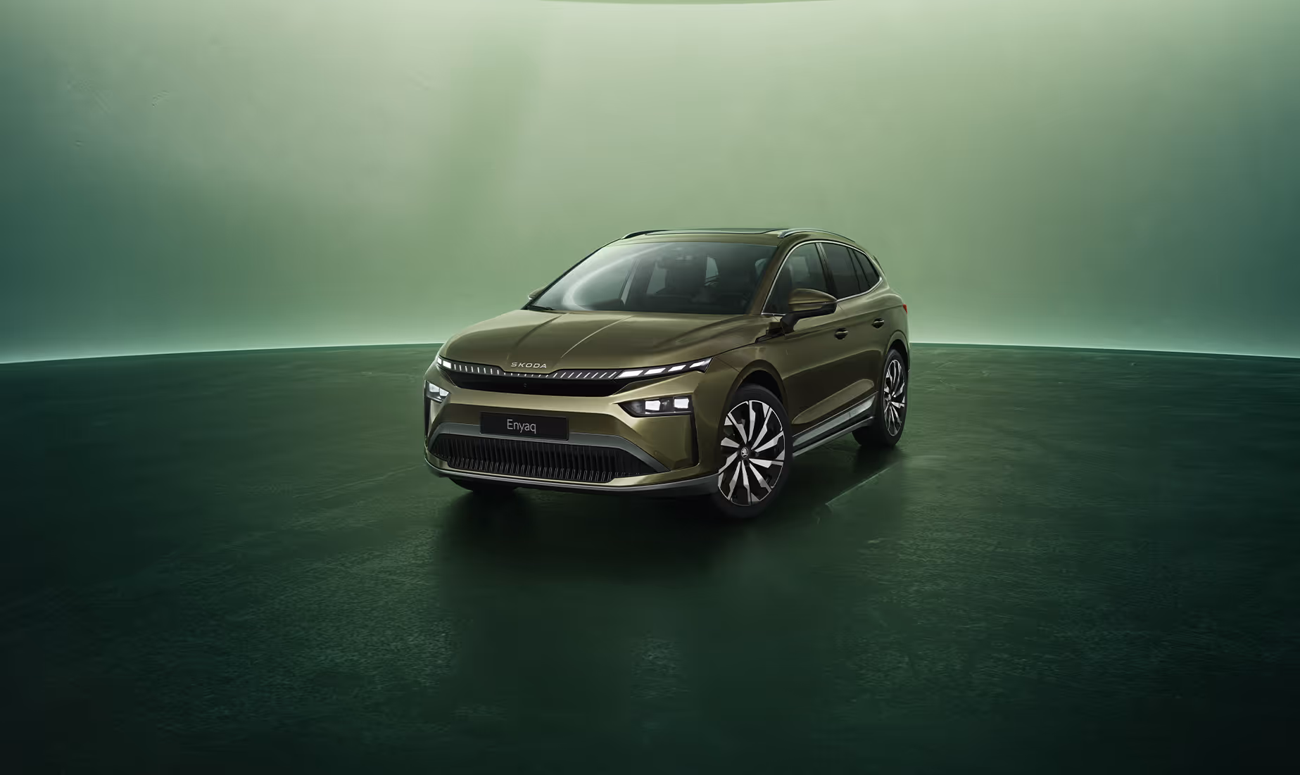 Dreiviertel-Frontansicht auf einen Škoda Enyaq in der Farbe Olibo-Grün Metallic vor einem grünen Studiohintergrund. Oben links steht geschrieben "Škoda 130 Jahre". Unten rechts ist der Schriftzug "Enyaq" platziert.