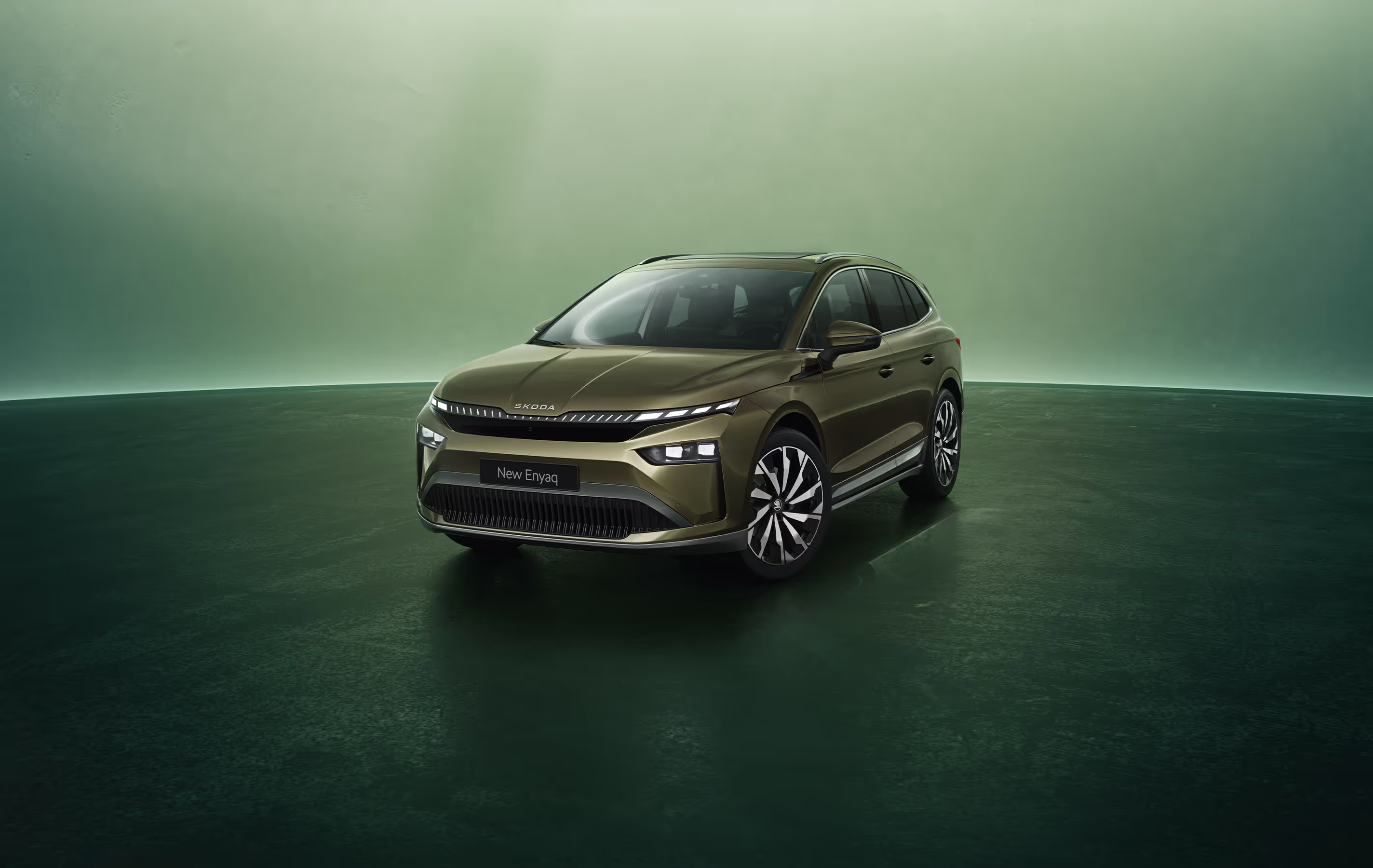 Dreiviertel-Frontansicht auf einen Škoda Enyaq in der Farbe Olibo-Grün Metallic vor einem grünen Studiohintergrund. Oben links steht geschrieben "Škoda 130 Jahre". Unten rechts ist der Schriftzug "Enyaq" platziert.