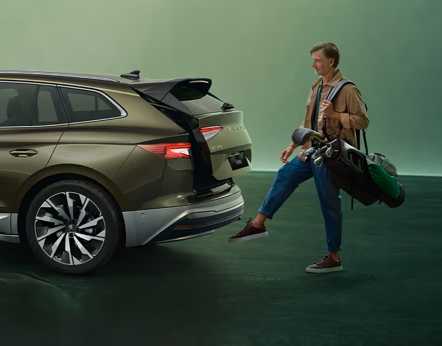 Ein Mann öffnete berührungslos den Kofferraum eines Škoda Elroq in der Farbe Olibo-Grün Metallic, indem er einen Fuß unter den Sensor hält. Die Person trägt eine Golftasche über der linken Schulter.