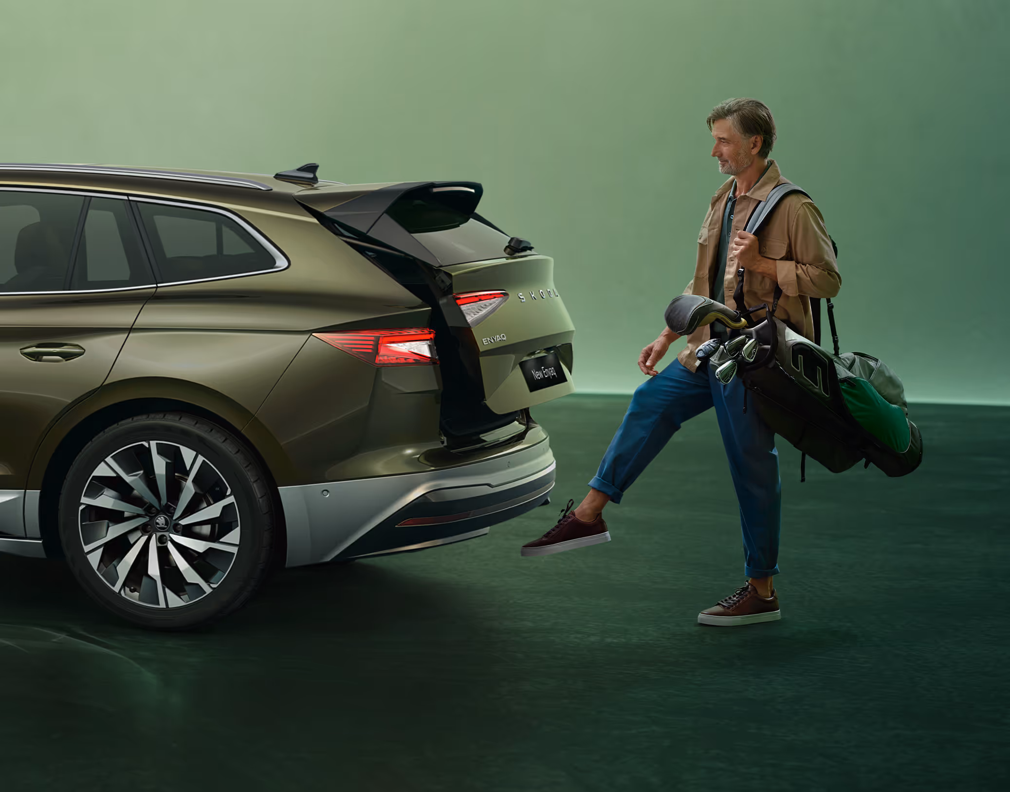 Ein Mann öffnete berührungslos den Kofferraum eines Škoda Elroq in der Farbe Olibo-Grün Metallic, indem er einen Fuß unter den Sensor hält. Die Person trägt eine Golftasche über der linken Schulter.
