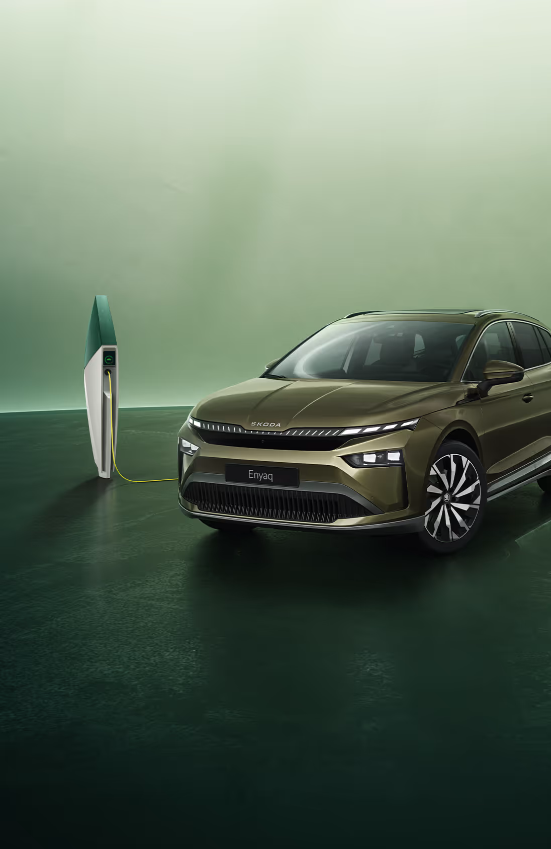 Schräge Frontansicht eines Škoda Enyaq in der Lackierung Olibo-Grün Metallic vor grünem Studiohintergrund. Das Fahrzeug ist über ein gelbes Kabel mit einer links im Bild stehenden futuristisch anmutenden Ladesäule verbunden.