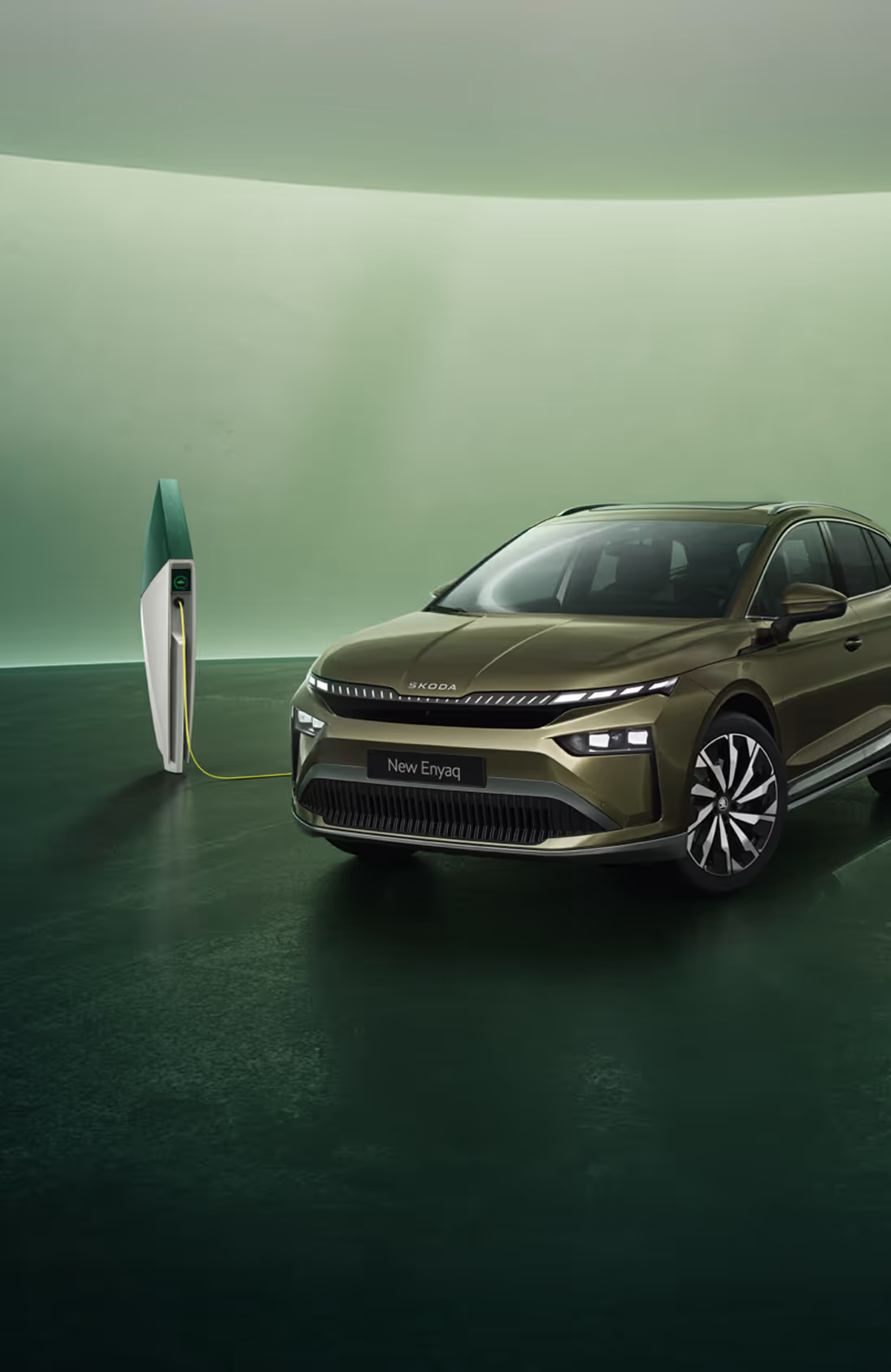 Schräge Frontansicht eines Škoda Enyaq in der Lackierung Olibo-Grün Metallic vor grünem Studiohintergrund. Das Fahrzeug ist über ein gelbes Kabel mit einer links im Bild stehenden futuristisch anmutenden Ladesäule verbunden.