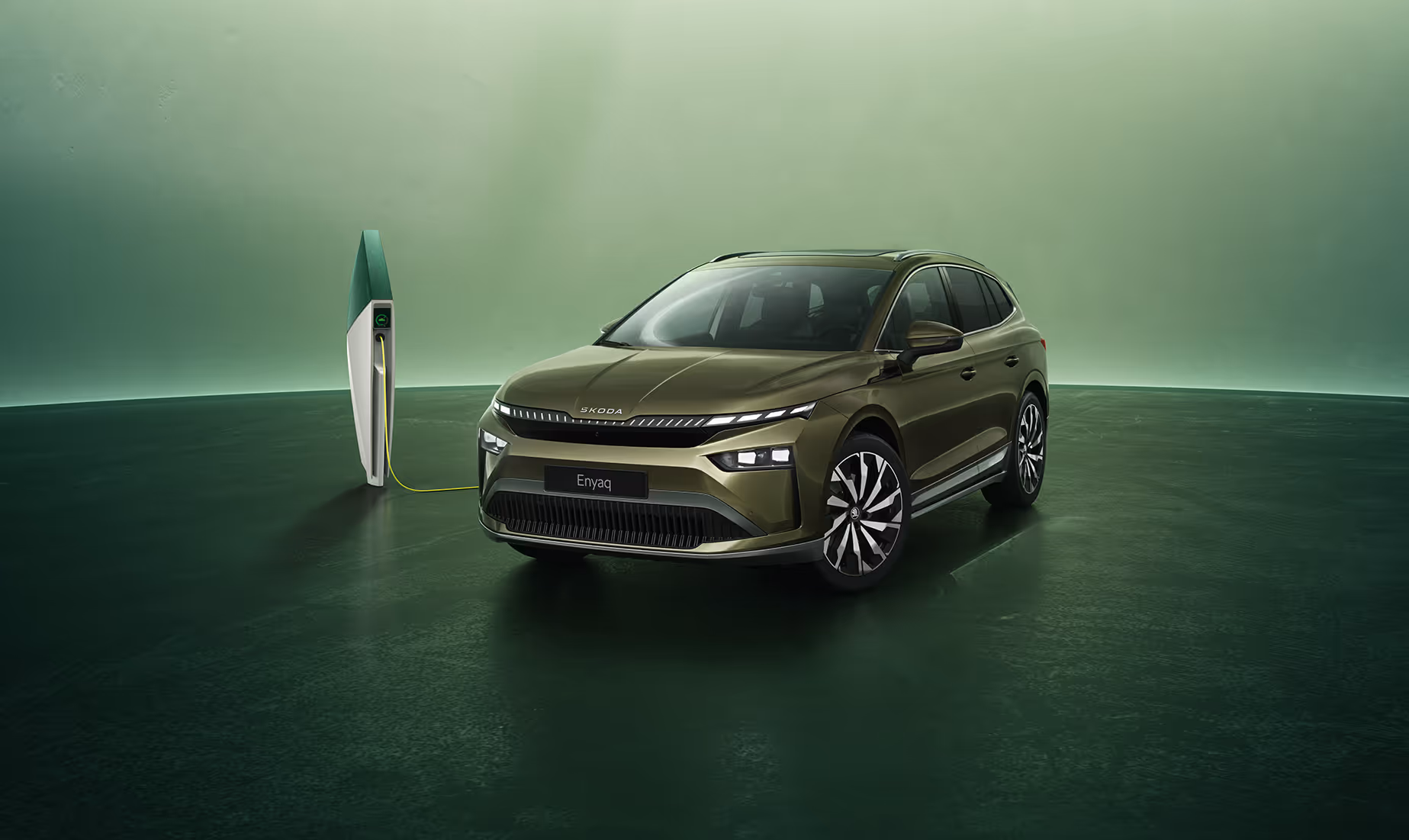 Schräge Frontansicht eines Škoda Enyaq in der Lackierung Olibo-Grün Metallic vor grünem Studiohintergrund. Das Fahrzeug ist über ein gelbes Kabel mit einer links im Bild stehenden futuristisch anmutenden Ladesäule verbunden.