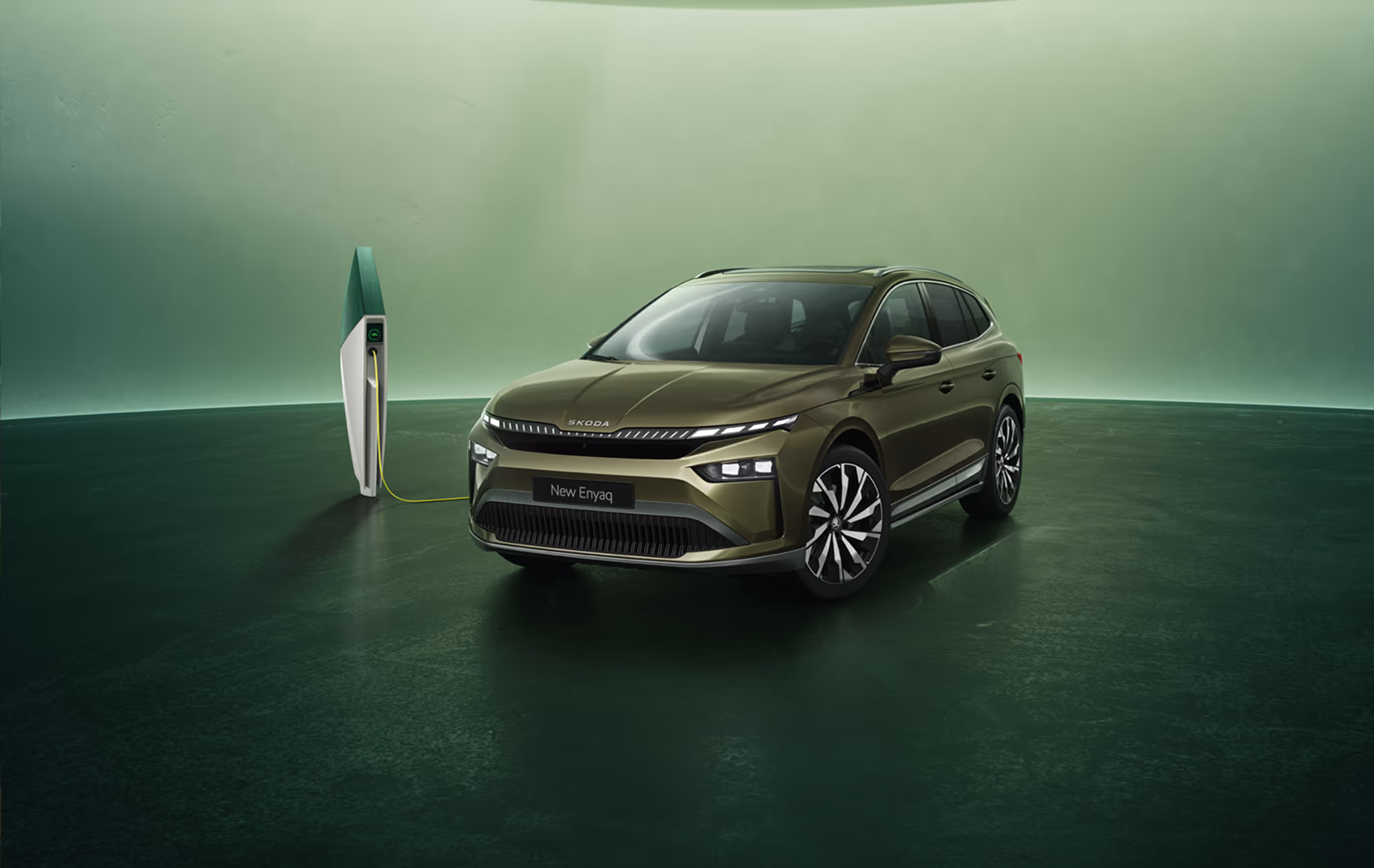 Schräge Frontansicht eines Škoda Enyaq in der Lackierung Olibo-Grün Metallic vor grünem Studiohintergrund. Das Fahrzeug ist über ein gelbes Kabel mit einer links im Bild stehenden futuristisch anmutenden Ladesäule verbunden.