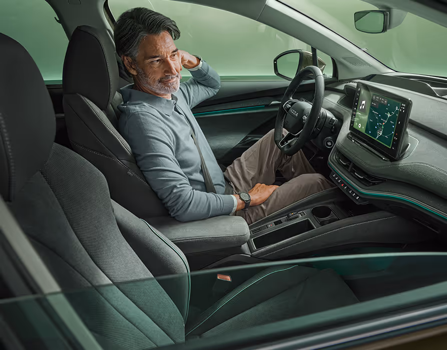 Blick durch das heruntergefahrene Beifahrerfenster ins Innere des Škoda Enyaq. Ein Mann sitzt auf dem Fahrersitz und blickt lächelnd auf das Infotainment-Display. Das Interieur ist mit dunklen Stoffen und einer grünen Ambientebeleuchtung ausgestattet.