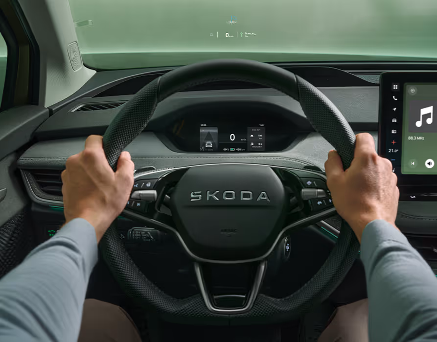 Aus der Fahrerperspektive sehen wir zwei Hände, die das Lenkrad eines Škoda Enyaq umfassen. Im Fokus stehen das Digital Cockpit hinter dem Lenkrad und das Head-up-Display, das Fahrinformationen direkt auf die Windschutzscheibe projiziert. Das Cockpit zeigt eine Geschwindigkeit von 0 km/h und eine Reichweite von 450 km an.