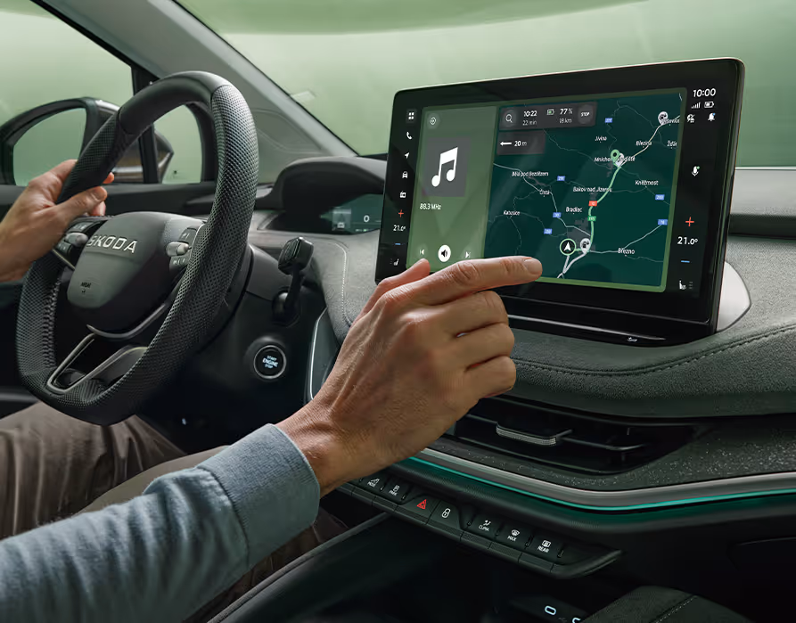 Im Innenraum eines Škoda Enyaq bedient eine Person mit dem Finger das freistehende 13-Zoll-Infotainmentdisplay. Auf dem Bildschirm ist eine Navigationskarte mit einer markierten Route zu sehen, während die andere Hand am Lenkrad liegt. Das moderne Cockpit wird durch eine grüne Ambientebeleuchtung akzentuiert.