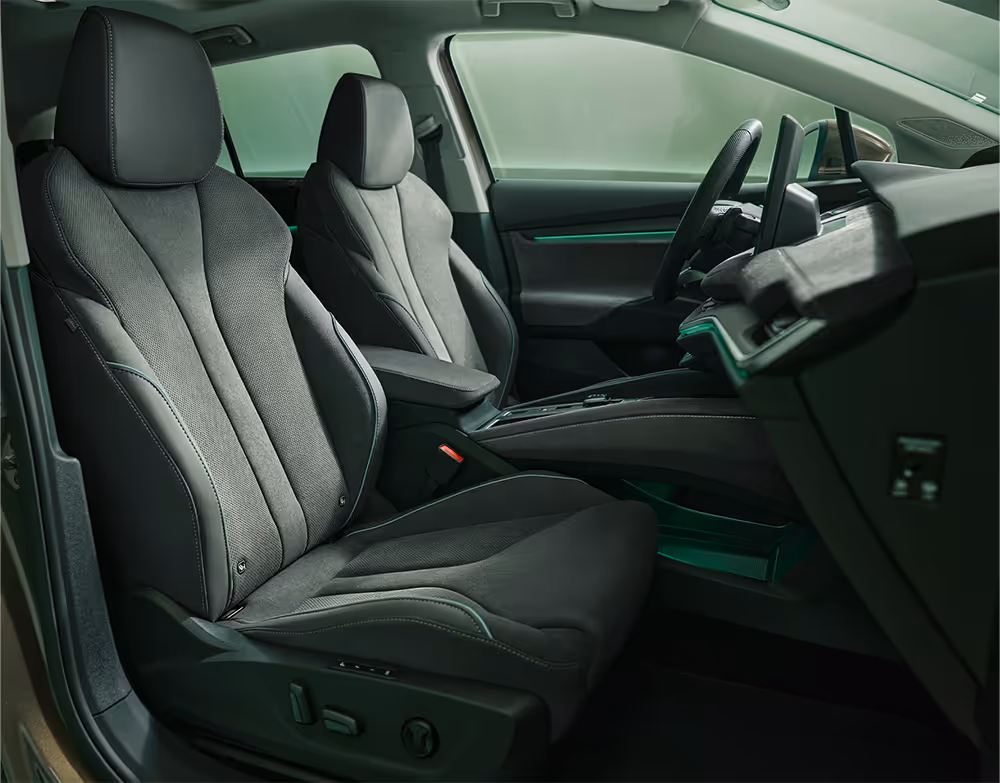 Ein Blick in den vorderen Innenraum des Škoda Enyaq, der die Fahrer- und Beifahrersitze in den Mittelpunkt stellt. Die Sitze der Design Selection „Lodge“ sind mit einer Kombination aus Stoff und Mikrofaser in dunklen Grautönen bezogen und weisen grüne Ziernähte auf. Die grüne Ambientebeleuchtung leuchtet entlang des Armaturenbretts und der Fahrertür.