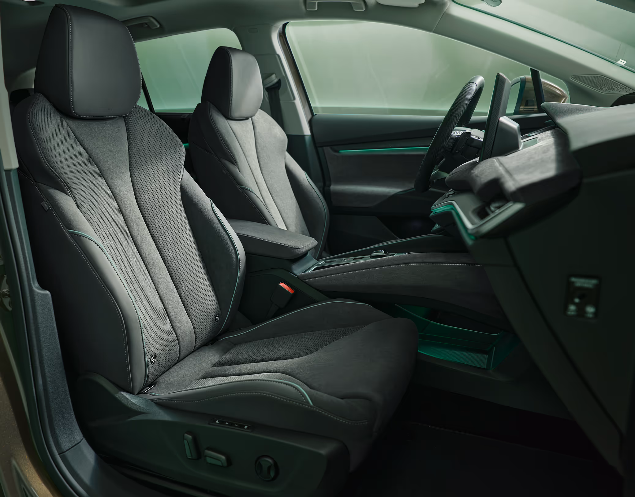 Ein Blick in den vorderen Innenraum des Škoda Enyaq, der die Fahrer- und Beifahrersitze in den Mittelpunkt stellt. Die Sitze der Design Selection „Lodge“ sind mit einer Kombination aus Stoff und Mikrofaser in dunklen Grautönen bezogen und weisen grüne Ziernähte auf. Die grüne Ambientebeleuchtung leuchtet entlang des Armaturenbretts und der Fahrertür.