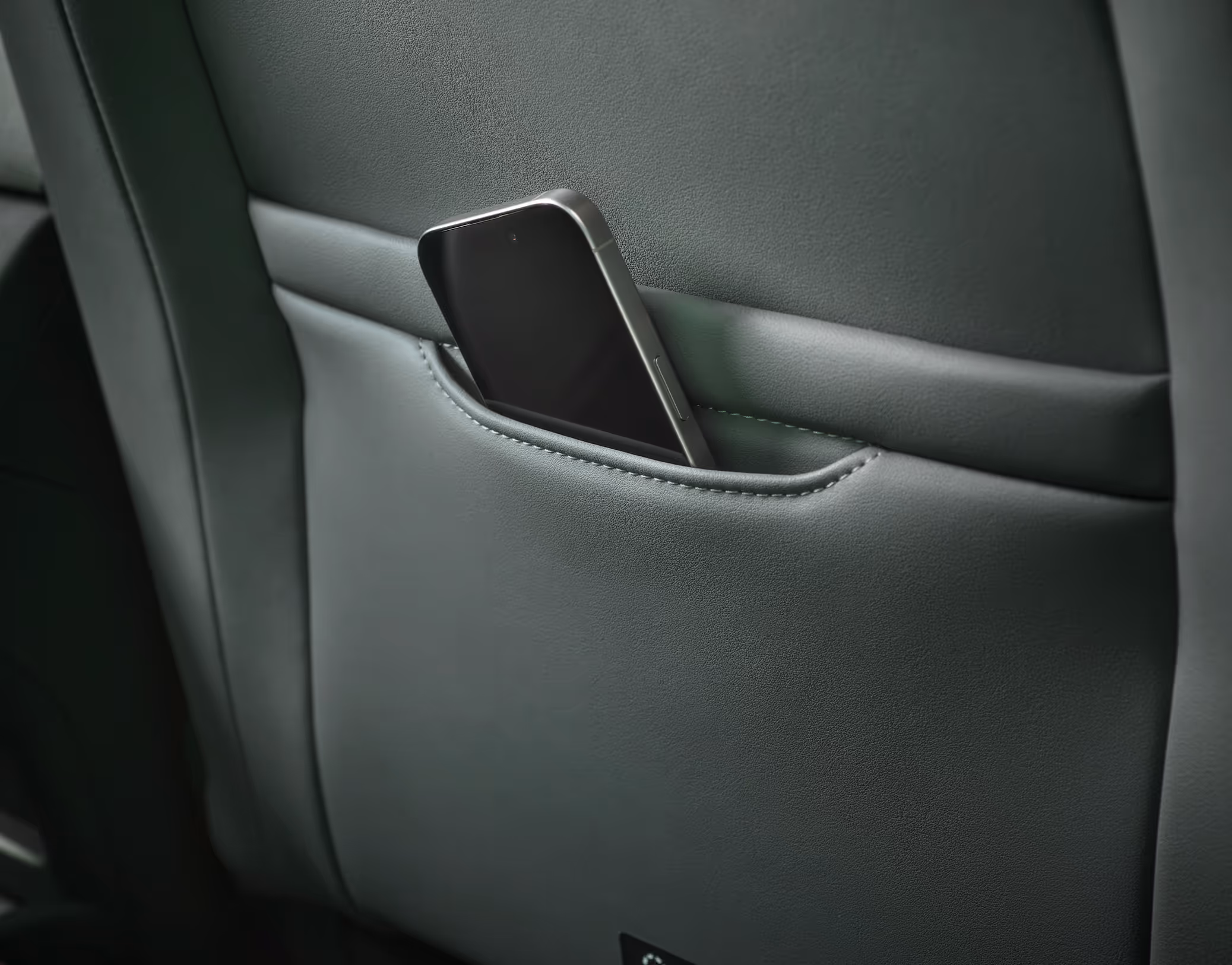 Eine Nahaufnahme zeigt die Rückseite eines Vordersitzes mit schwarzem Lederbezug im Innenraum eines Škoda Enyaq. In einer kleinen, aufgesetzten Tasche steckt ein Smartphone. Links im unscharfen Hintergrund sind die Lüftungsdüsen der Mittelkonsole für den Fondbereich zu erkennen.