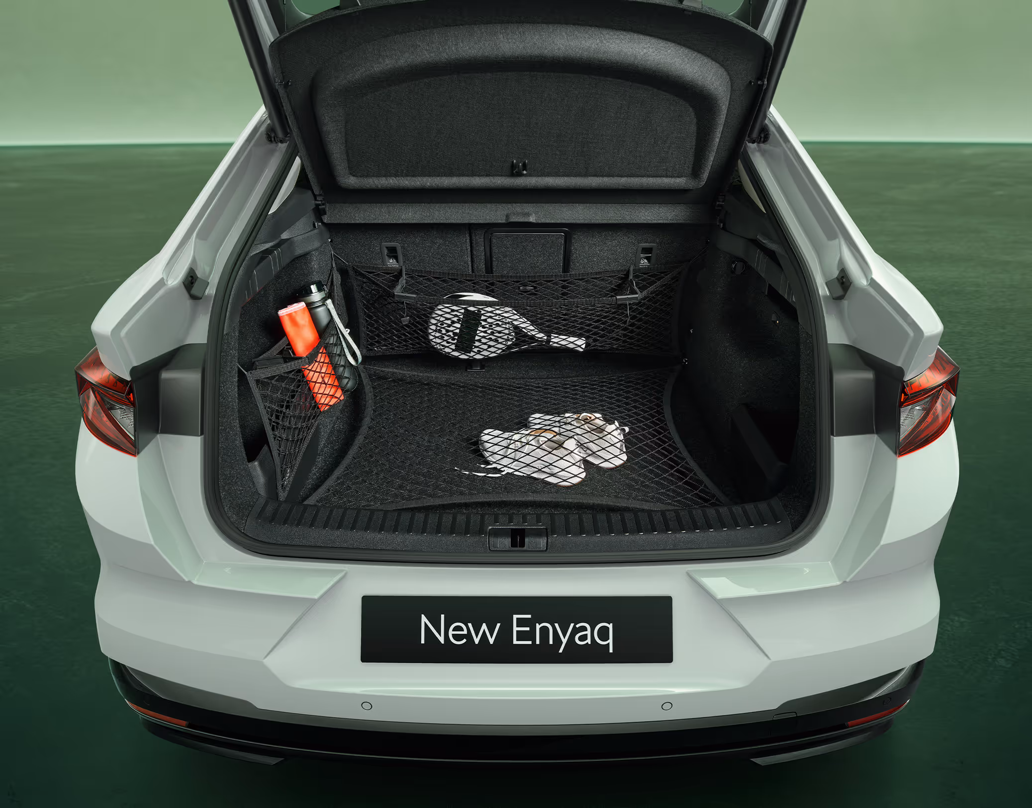 Blick in den geöffneten, leeren Kofferraum des Škoda Enyaq Coupé in der Farbe Moon-Weiß Perleffekt vor grünem Studiohintergrund. Das „Simply Clever“-Netzprogramm ist im Einsatz und sichert verschiedene Gegenstände wie einen Paddelschläger, Turnschuhe und eine Trinkflasche.