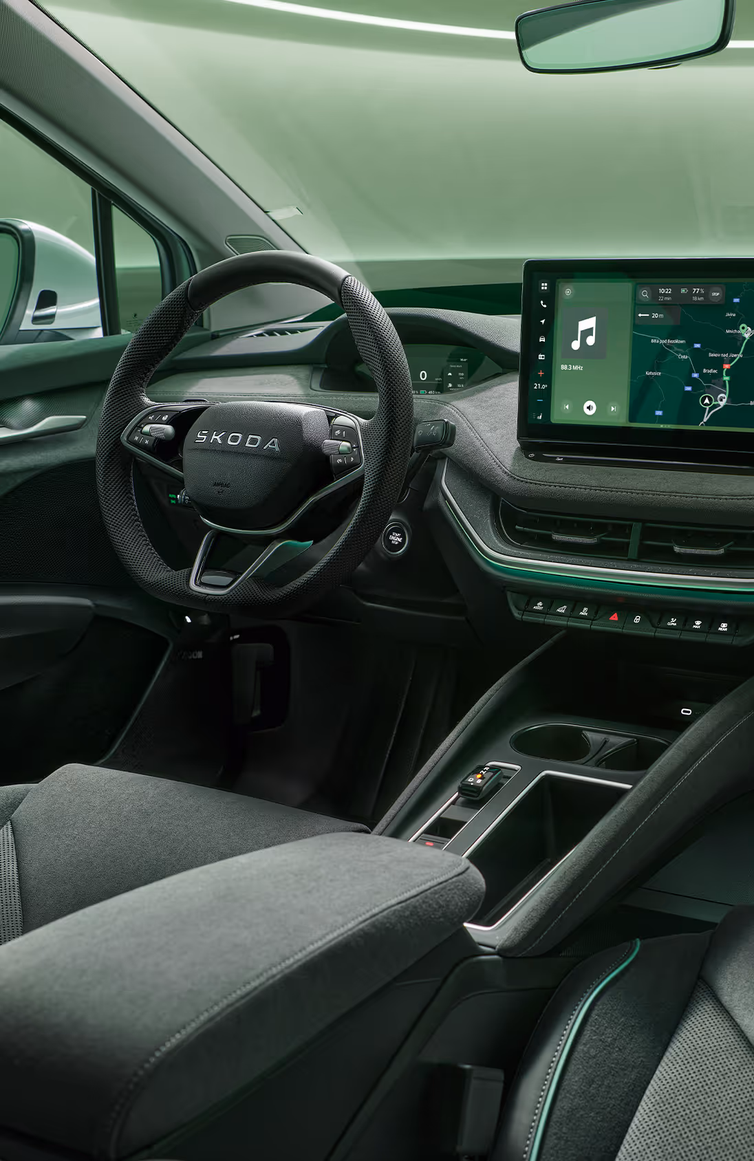 Fokus auf das Armaturenbrett und das Infotainmentsystem des Škoda Enyaq. Zentral platziert ist das freistehende 13-Zoll-Infotainmentdisplay, das eine Navigationskarte und diverse Fahrzeugfunktionen anzeigt. Das dunkle Interieur der Design Selection „Lodge“ wird durch eine grüne Ambientebeleuchtung abgerundet. Unten rechts steht auf einer dunkelgrün abgesetzten Fläche der Schriftzug "Škoda Connect".