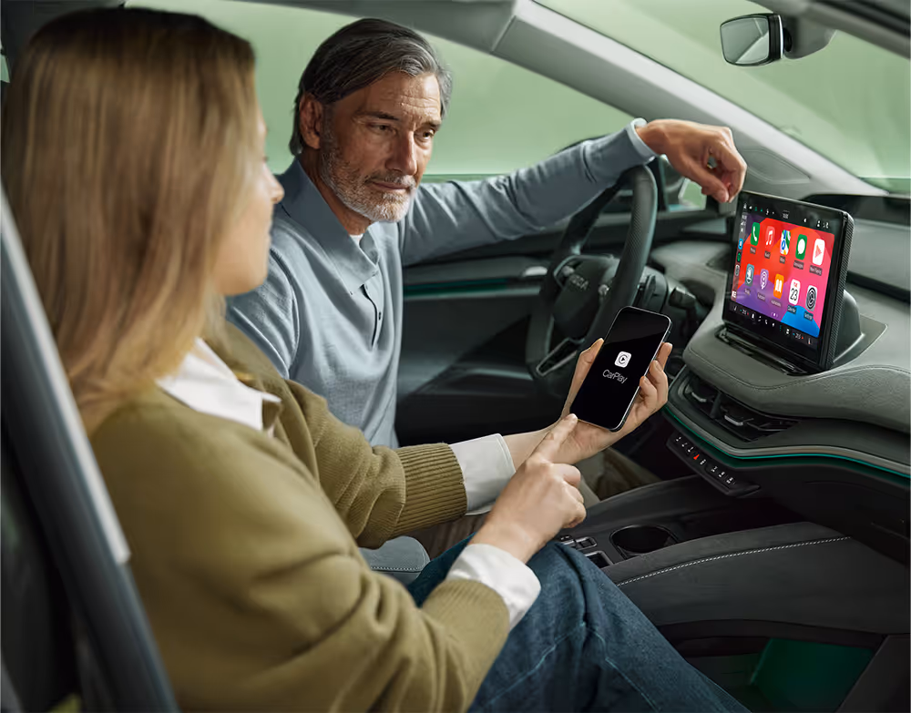 Eine Frau auf dem Beifahrersitz eines Škoda Enyaq verbindet ihr Smartphone über Wireless SmartLink mit dem Fahrzeug. Auf dem Smartphone ist das Apple CarPlay-Logo zu sehen, während die Benutzeroberfläche mit den Apps bereits auf dem großen 13-Zoll-Infotainmentdisplay gespiegelt wird. Der Mann am Steuer blickt interessiert auf das Display.
