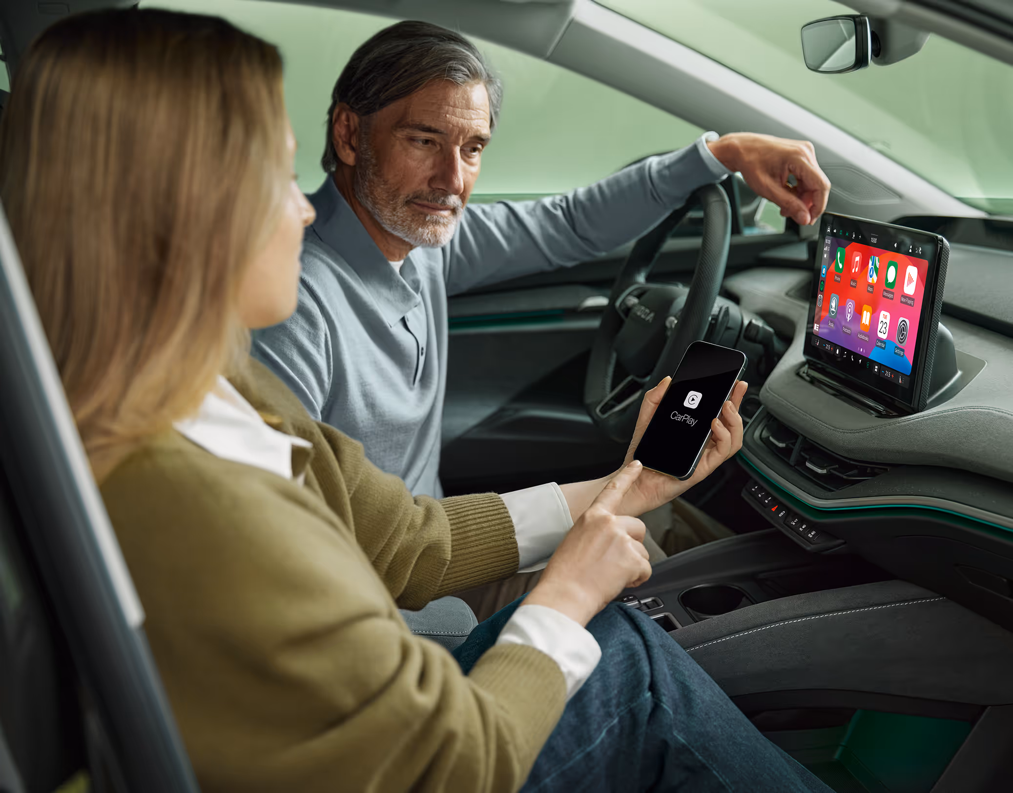 Eine Frau auf dem Beifahrersitz eines Škoda Enyaq verbindet ihr Smartphone über Wireless SmartLink mit dem Fahrzeug. Auf dem Smartphone ist das Apple CarPlay-Logo zu sehen, während die Benutzeroberfläche mit den Apps bereits auf dem großen 13-Zoll-Infotainmentdisplay gespiegelt wird. Der Mann am Steuer blickt interessiert auf das Display.