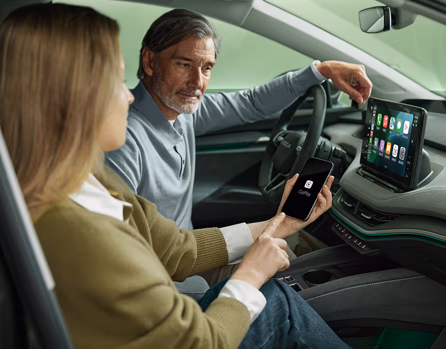 Eine Frau auf dem Beifahrersitz eines Škoda Enyaq verbindet ihr Smartphone über Wireless SmartLink mit dem Fahrzeug. Auf dem Smartphone ist das Apple CarPlay-Logo zu sehen, während die Benutzeroberfläche mit den Apps bereits auf dem großen 13-Zoll-Infotainmentdisplay gespiegelt wird. Der Mann am Steuer blickt interessiert auf das Display.